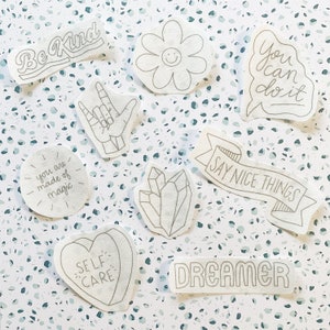 Puede incluir: Un conjunto de diez parches termoadhesivos blancos con varios mensajes y diseños positivos, que incluyen "Sé amable", "Estás hecho de magia", "Puedes hacerlo", "Di cosas agradables", "Soñador", "Cuídate", una mano haciendo el signo de "Te amo", un cristal y una flor.