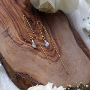 Könnte beinhalten: Ein Paar goldfarbene Ohrringe mit tropfenförmigen, schillernden Edelsteinen. Die Ohrringe werden auf einer Holzoberfläche präsentiert, mit weißen Blumen und Grünzeug im Hintergrund.