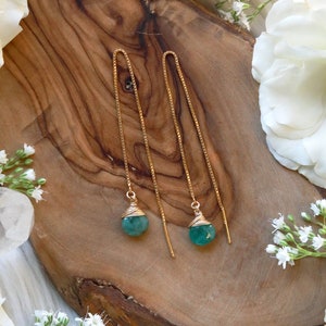May include: Gold-toned threader earrings with teardrop-shaped, teal-colored gemstones. The gemstones are wire-wrapped and suspended from delicate chains. The earrings rest on a wooden surface, surrounded by white flowers.
