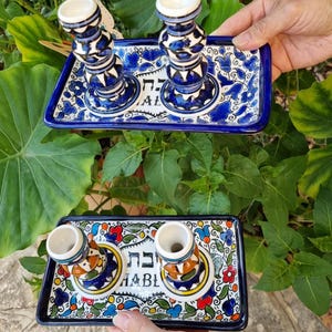 May include: Two ceramic candle holders on a rectangular tray. The candle holders are blue and white with a floral design. The tray is also blue and white with a floral pattern and Hebrew text. The second tray has a colorful floral design.