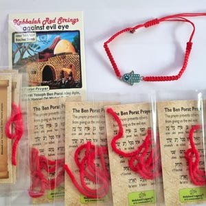 May include: A red string bracelet featuring a small hamsa charm with a blue eye detail, accompanied by several prayer cards. The cards include text titled Kabbalah Red Strings against evil eye, Prayer for Traveler, and The Ben Porat Prayer.