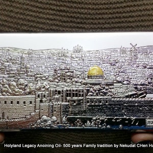 May include: A silver metal box with a detailed image of the city of Jerusalem. The box has a gold dome on top and the word "Jerusalem" printed on the bottom. The text "Holyland Legacy Anointing Oil - 500 years Family tradition by Nekudat Chen Haifa 972-546818597" is printed below the box.