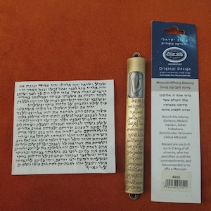 Op de afbeelding: Een goudkleurige mezuzah-koker met Hebreeuwse tekst, een wit papier met Hebreeuwse tekst en een kaart met Engelse en Hebreeuwse tekst. De mezuzah-koker is cilindrisch met een lus aan de bovenkant. De kaart leest "Mezuzah Affixing Blessing".