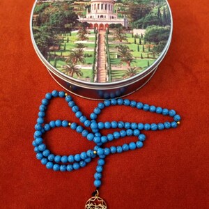 May include: A round tin featuring a picture of a building with a golden dome and a long pathway. A blue beaded necklace with a gold pendant is in front of the tin. The background is a solid red colour.
