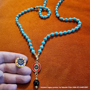 May include: A turquoise blue beaded necklace with a gold chain and a red and gold pendant. The pendant has a black and gold design. The necklace is on a red surface.