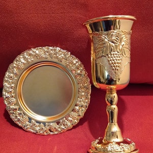 Könnte beinhalten: Ein goldfarbenes Kelch- und Teller-Set. Der Kelch hat Trauben- und Blattmotive, und der Teller hat einen dekorativen Trauben- und Blattrand. Das Set wird auf einer roten Oberfläche präsentiert.