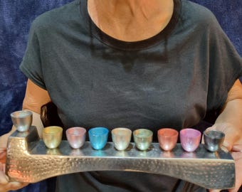 May include: A silver menorah with nine cups, each with a different iridescent color. The cups are arranged in a row on a textured base.