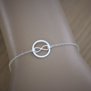 Peut inclure: Un bracelet argenté orné d'un symbole infini dans un cercle. Le bracelet est composé de maillons de chaîne fins. Le symbole infini est en forme de huit. Le bracelet est présenté sur un poignet.