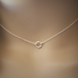 May include: A delicate silver necklace featuring a small, open circle pendant. The necklace is displayed against a neutral background, with a denim jacket visible on the side. The chain is fine and the pendant is centered.