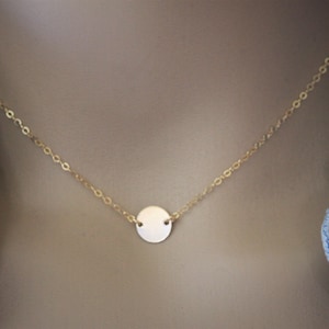 May include: A delicate gold-colored necklace featuring a small, round pendant. The pendant is centered on a fine chain, creating a minimalist and elegant design. The necklace is shown against a neutral background, highlighting its simplicity.