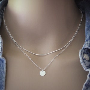 Puede incluir: Un collar de plata con una cadena delicada y un pequeño colgante redondo. El collar está superpuesto con una segunda cadena más fina.