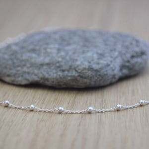 Op de afbeelding: Een delicate zilveren ketting met kleine, gelijkmatig verdeelde kralen. De ketting wordt getoond op een lichtbruin houten oppervlak, met een grijze steen op de achtergrond. De ketting is een eenvoudig, elegant sieraad.