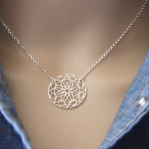 Könnte beinhalten: Eine silberne Halskette mit einem zarten, runden Anhänger mit geometrischem Muster. Der Anhänger besteht aus filigranem, durchbrochenem Metall und ähnelt einem Mandala.