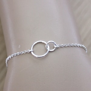 Puede incluir: Un brazalete de cadena de plata con dos círculos entrelazados como pieza central.
