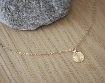 Può includere: Una delicata collana color oro con un piccolo pendente rotondo. Il pendente presenta un motivo a raggi di sole strutturato. La collana è esposta su una superficie di legno, con una pietra grigia sullo sfondo.