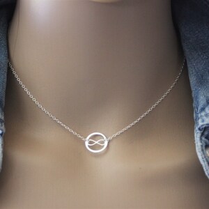 Könnte beinhalten: Eine zarte Silberkette mit einem kreisförmigen Anhänger mit einem Unendlichkeitssymbol in der Mitte. Die Kette ist fein und der Anhänger ist zentriert. Die Halskette wird vor einer Jeansjacke präsentiert.