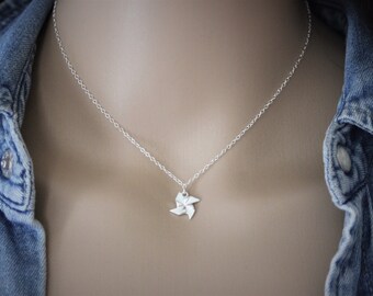 May include: A silver-colored necklace with a small pinwheel pendant. The pendant is a detailed, three-dimensional design. The necklace is displayed against a neutral background, with a denim jacket visible in the frame.
