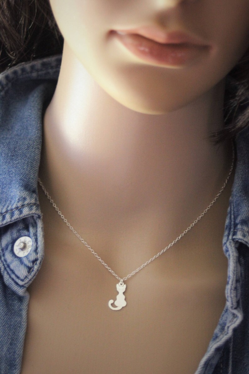K&ouml;nnte beinhalten: Eine silberne Kettenkette mit einem kleinen, silbernen Katzenanh&auml;nger. Die Kette wird von einer Person mit einer Jeansjacke getragen.