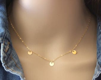 May include: A delicate gold-colored necklace featuring three small, round pendants. The necklace is displayed against a denim jacket, highlighting the jewelry's simple elegance. The chain and pendants are a warm, shiny gold.