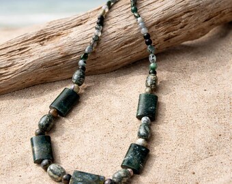 May include: A beaded necklace featuring a mix of dark green cylindrical stone beads and smaller round mottled green, gray, and black stone beads. The necklace is secured with a silver-toned metal clasp at the top, showcasing a variety of natural stone textures and earthy color tones throughout the piece.