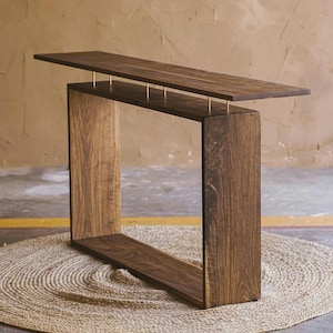 May include: A modern, dark wood console table with a rectangular frame and a floating top. The top is supported by thin, gold-colored metal rods. The table is placed on a round, woven jute rug.