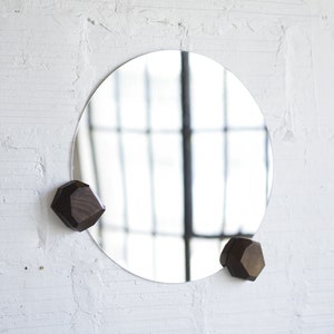 May include: A round mirror with a wooden frame hangs on a white brick wall. Two dark brown wooden geometric hooks are attached to the wall on either side of the mirror.