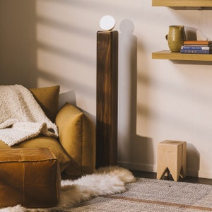 May include: A tall, dark wooden lamp with a round, white light at the top stands in a room with a mustard-colored sofa, a leather ottoman, and a small wooden stool. A white fur rug and a patterned rug are on the floor. Shelves with books and a vase are on the wall.