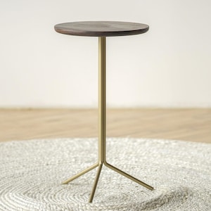 May include: A tall, slender side table featuring a circular dark brown wooden top supported by a single vertical post that branches into a three-legged base made of smooth, matte gold-toned metal, providing a stable foundation for the elevated surface.