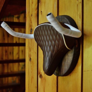 May include: A decorative wall mount featuring a brown bicycle seat with white trim and white handlebar tape. The seat is mounted on a dark wooden plaque and is displayed against a wooden wall.