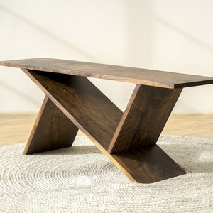 May include: A modern, dark wood table with a unique X-shaped base. The tabletop is rectangular and the legs are angled, creating a contemporary design. The table is set on a round, light-colored rug.