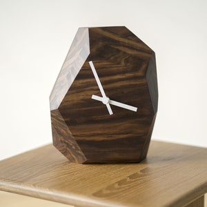 May include: A dark brown wooden clock with a geometric shape and white hands. The clock is sitting on a light brown wooden table.