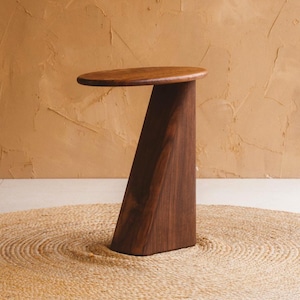 May include: A dark brown wooden side table with a round top and a triangular base. The table is set on a round, woven jute rug. The background is a textured, tan wall.