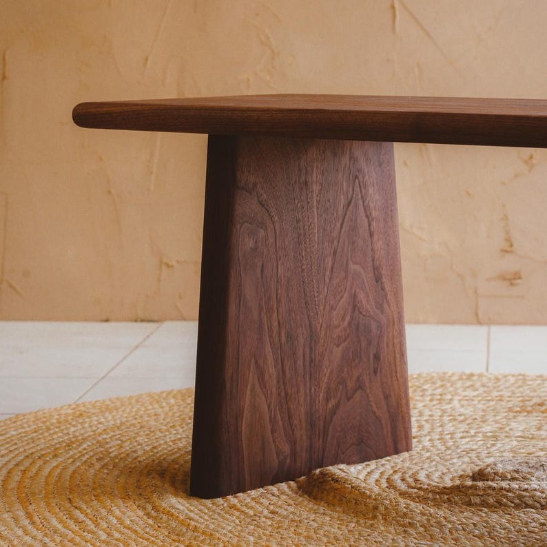 Puede incluir: Un banco de madera de color marr&oacute;n oscuro con una superficie superior horizontal lisa y una &uacute;nica pata de soporte vertical ancha y c&oacute;nica. La veta de la madera es claramente visible, mostrando patrones naturales y un rico color marr&oacute;n intenso.
