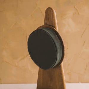 K&ouml;nnte beinhalten: Ein dunkelolivgr&uuml;nes, kreisf&ouml;rmiges Kissen mit glatter, matter Textur ist auf einem hohen, hellbraunen Holzst&auml;nder montiert. Der St&auml;nder hat ein vertikales, spitz zulaufendes Design mit einem runden Rahmen, der das Kissen sicher h&auml;lt.