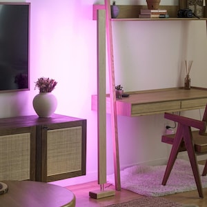 May include: A modern interior scene with a wooden desk, chair, and cabinet. A tall, light-colored wooden floor lamp stands next to a wall. The room is illuminated with a pinkish-purple light, creating a warm and inviting atmosphere.