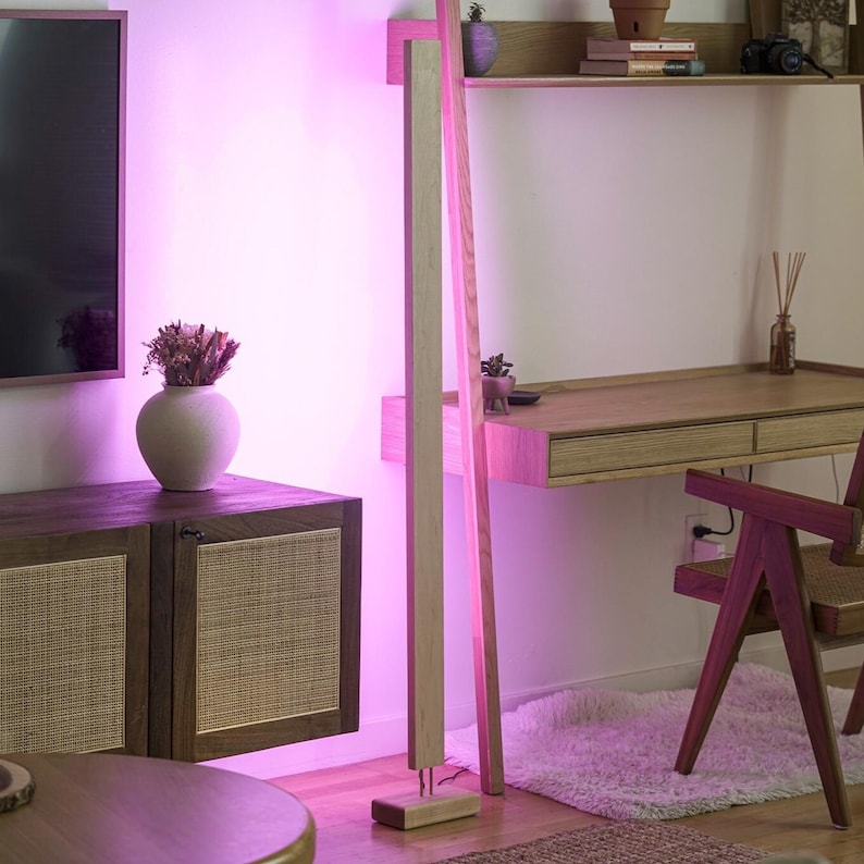 May include: A modern interior scene with a wooden desk, chair, and cabinet. A tall, light-colored wooden floor lamp stands next to a wall. The room is illuminated with a pinkish-purple light, creating a warm and inviting atmosphere.