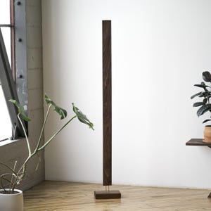 May include: A tall, dark brown wooden floor lamp with a rectangular base and a slender, vertical design. The lamp stands on a light-colored wooden floor, with a white wall in the background. A potted plant is visible on the left.