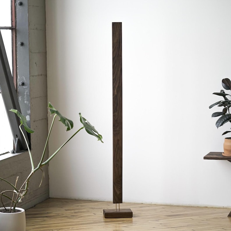 May include: A tall, dark brown wooden floor lamp with a rectangular base and a slender, vertical design. The lamp stands on a light-colored wooden floor, with a white wall in the background. A potted plant is visible on the left.