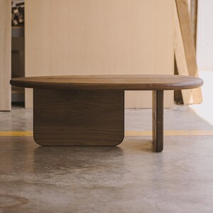 May include: A solid wood coffee table with a dark brown finish. The table has a rounded rectangular top and a unique base design with a rectangular support and a single leg. The table is made of a dark wood.