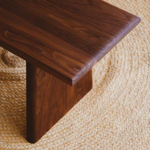 Puede incluir: Un banco de madera de color marr&oacute;n oscuro con una superficie lisa y pulida que muestra vetas naturales. El banco tiene una parte superior rectangular gruesa con bordes redondeados y una base de soporte vertical s&oacute;lida de madera.