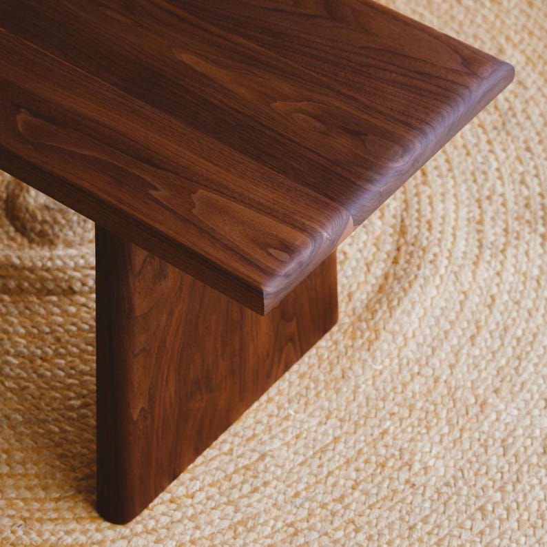 Puede incluir: Un banco de madera de color marr&oacute;n oscuro con una superficie lisa y pulida que muestra vetas naturales. El banco tiene una parte superior rectangular gruesa con bordes redondeados y una base de soporte vertical s&oacute;lida de madera.