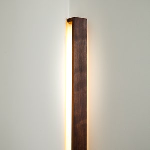 Puede incluir: Una l&aacute;mpara de pared vertical con un marco largo y rectangular de madera de color marr&oacute;n oscuro. El aplique est&aacute; instalado en una esquina y emite un brillo c&aacute;lido y suave desde detr&aacute;s de la madera, iluminando las paredes blancas.