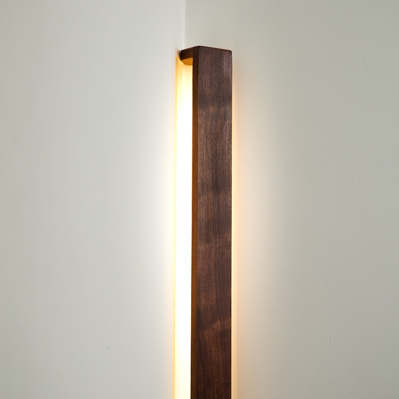 Puede incluir: Una l&aacute;mpara de pared vertical con un marco largo y rectangular de madera de color marr&oacute;n oscuro. El aplique est&aacute; instalado en una esquina y emite un brillo c&aacute;lido y suave desde detr&aacute;s de la madera, iluminando las paredes blancas.