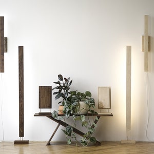 May include: Wooden wall and floor lamps with integrated LED lighting. The lamps are rectangular in shape and come in various wood tones, including dark brown and light beige. A small wooden table with a plant is in the center.