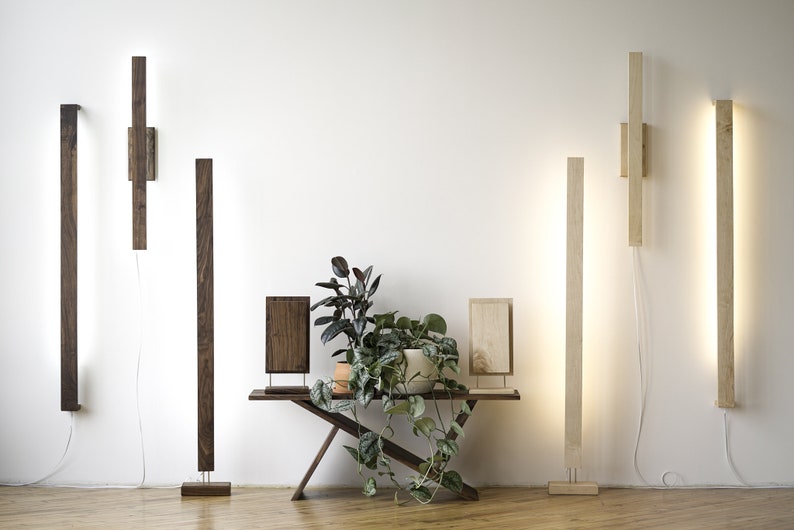 May include: Wooden wall and floor lamps with integrated LED lighting. The lamps are rectangular in shape and come in various wood tones, including dark brown and light beige. A small wooden table with a plant is in the center.
