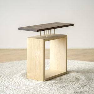 May include: A modern side table with a rectangular dark brown top supported by brass rods on a light wood base. The base is a light-colored, open-sided cube. The table sits on a round, textured, off-white rug.