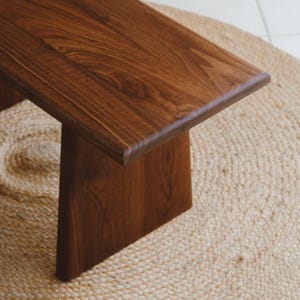 Puede incluir: Un banco de madera con una superficie lisa de color marr&oacute;n oscuro y un patr&oacute;n de veta natural visible. El banco est&aacute; construido con una parte superior rectangular plana y una pata de soporte s&oacute;lida e inclinada que se extiende hacia abajo.