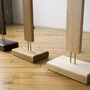 May include: Three wooden sculptures with rectangular bases and vertical posts. The sculptures are made of different types of wood, including dark brown, light brown, and natural wood. The posts are supported by brass rods.
