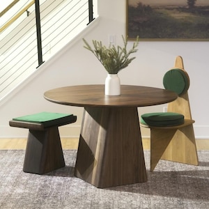 K&ouml;nnte beinhalten: Ein runder Esstisch aus dunklem Holz mit einem S&auml;ulenfu&szlig;, begleitet von einem passenden Hocker aus dunklem Holz und einem Stuhl aus hellem Holz, beide mit gr&uuml;nen Sitzkissen. Eine wei&szlig;e Vase mit gr&uuml;nen Kiefernzweigen steht auf dem Tisch.