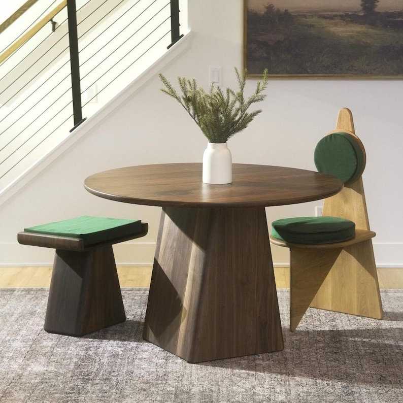 K&ouml;nnte beinhalten: Ein runder Esstisch aus dunklem Holz mit einem S&auml;ulenfu&szlig;, begleitet von einem passenden Hocker aus dunklem Holz und einem Stuhl aus hellem Holz, beide mit gr&uuml;nen Sitzkissen. Eine wei&szlig;e Vase mit gr&uuml;nen Kiefernzweigen steht auf dem Tisch.